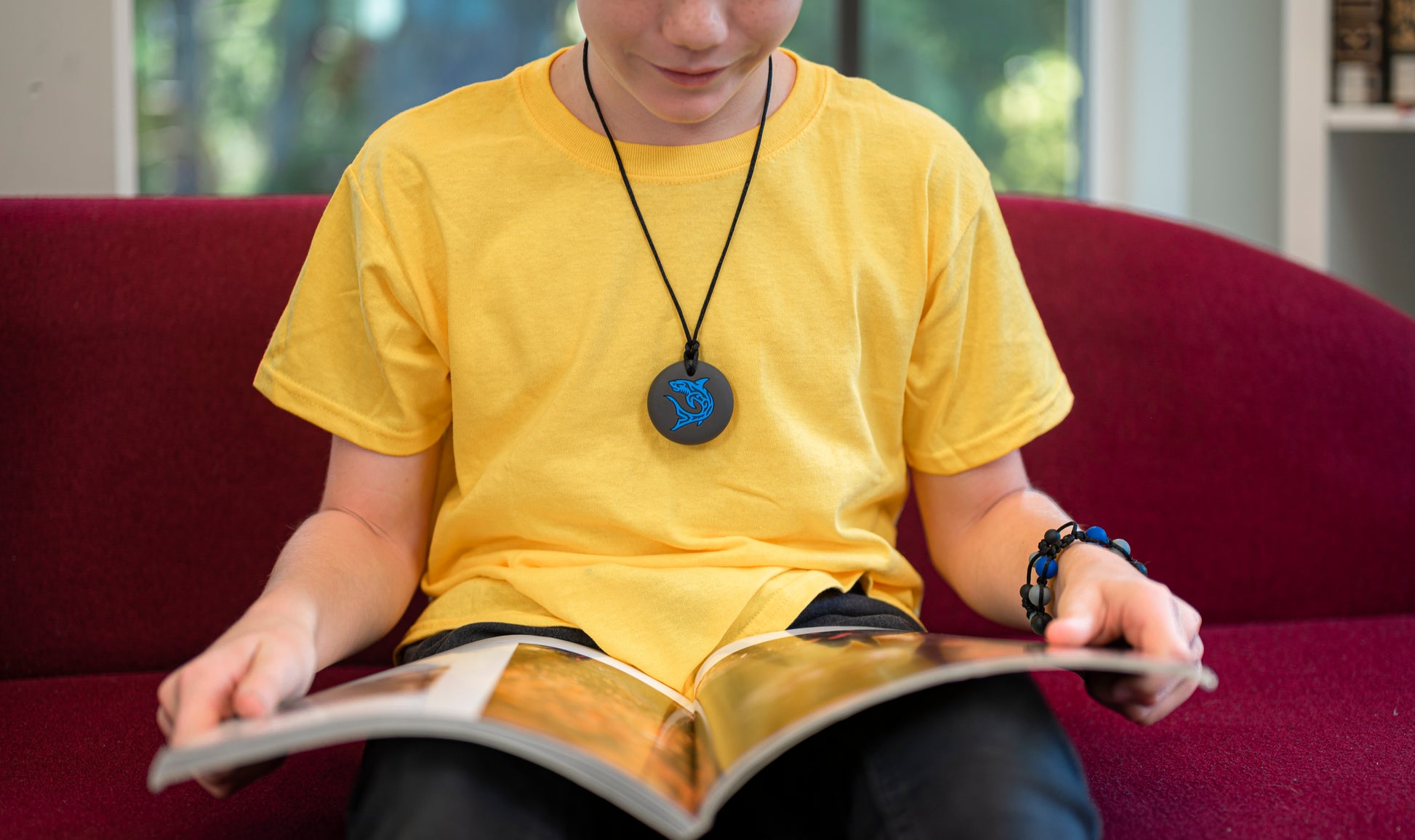 Boy reads while wearing Munchables Shark Chew Necklace in blue